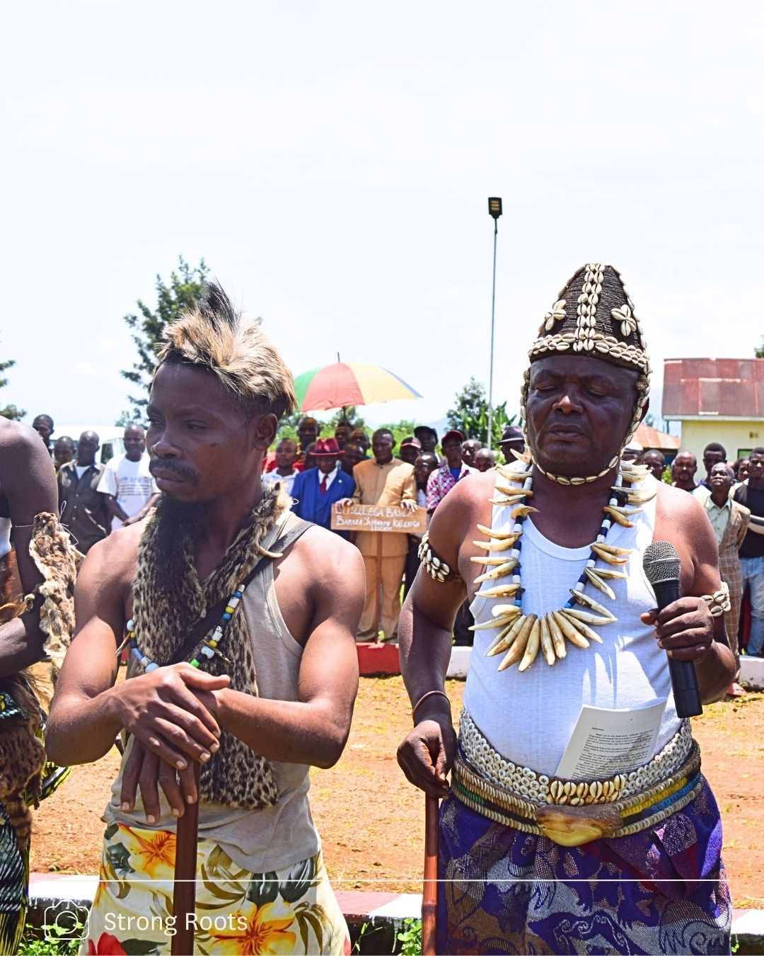 Two men dressed in traditional ceremonial regalia address a crowd at an outdoor event