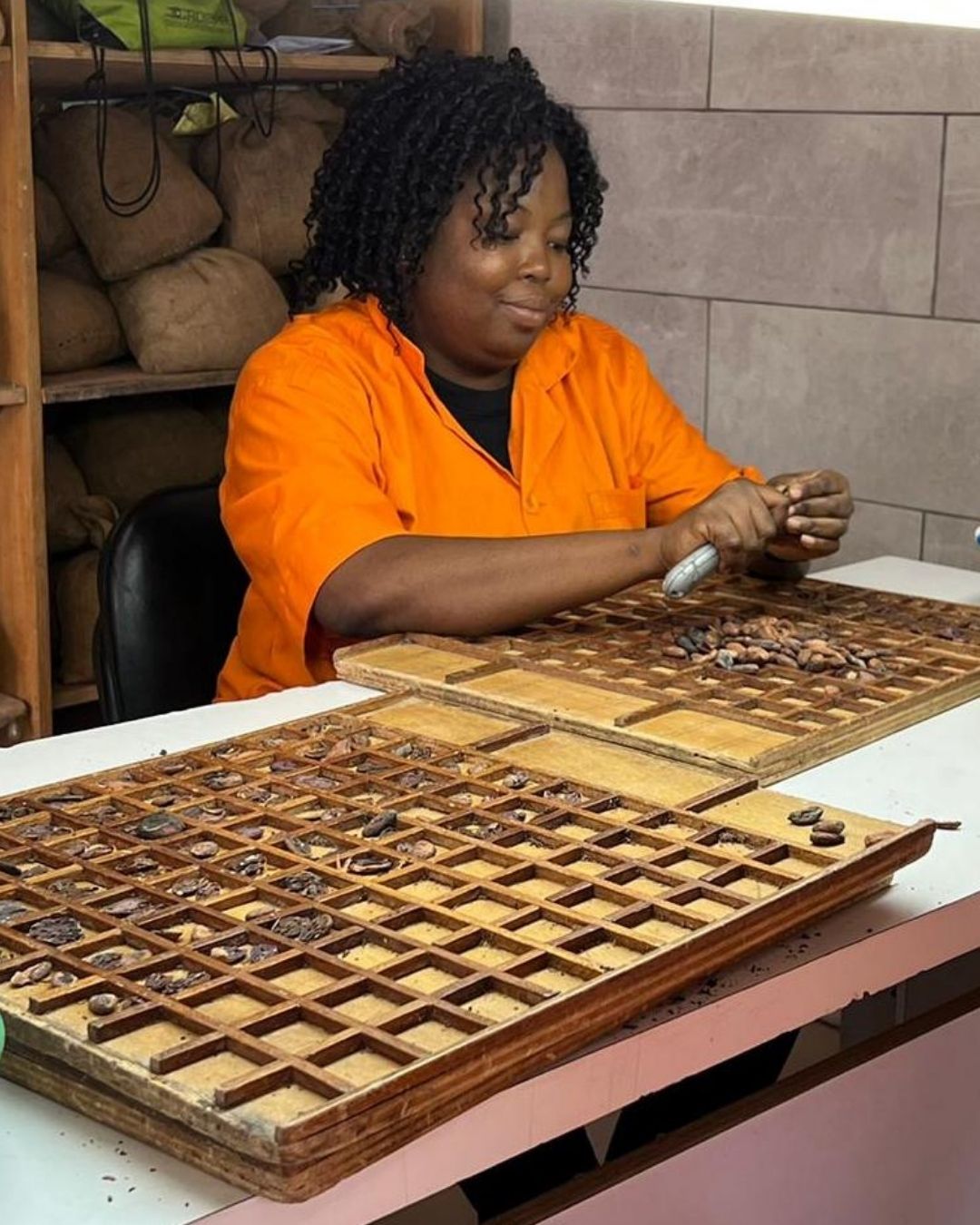 A woman in an orange shirt sorting cocoa beans into a sectioned wooden tray at a processing facility