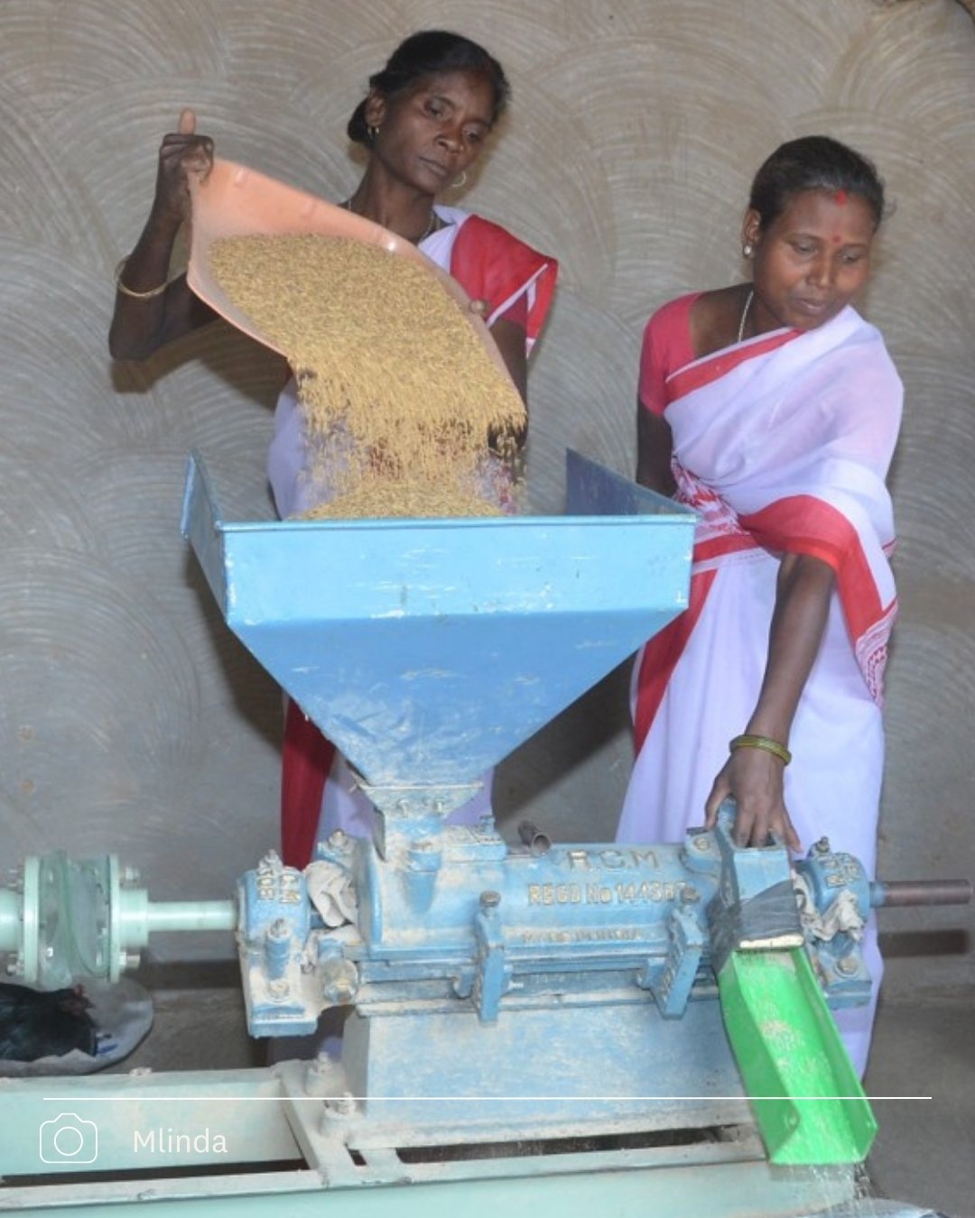 Two women in saris feeding grain into a blue mechanical grain mill